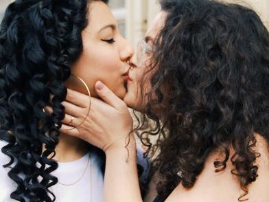 Two curly haired women kissing in front of a building.
