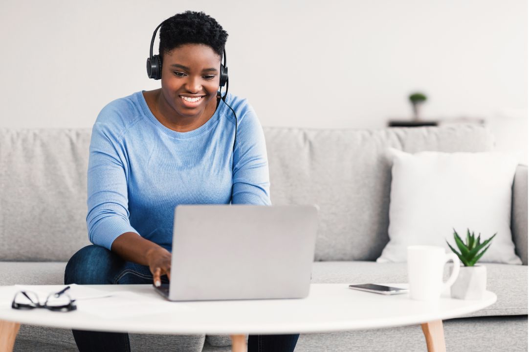 A woman wearing headphones is sitting on a couch and using a laptop.